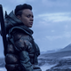 A woman (Leah Harvey's Salvor Hardin) wearing a parka with a weapon strapped to her back stands against a desolate backdrop in a scene from Foundation.
